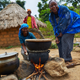 A woman laughing while she bends over a cookpot over an open fire. There are brick huts in the background with thatched roofs and three other people smiling in the picture.